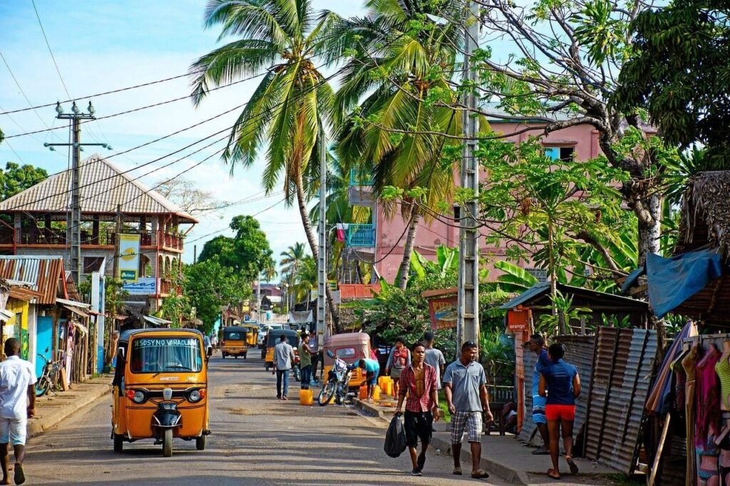 Belebte Strasse im Hauptort von Sainte Marie in Madagaskar mit gelben Tuk-Tuks, Fussgängern und Palmen zwischen bunten Häusern.