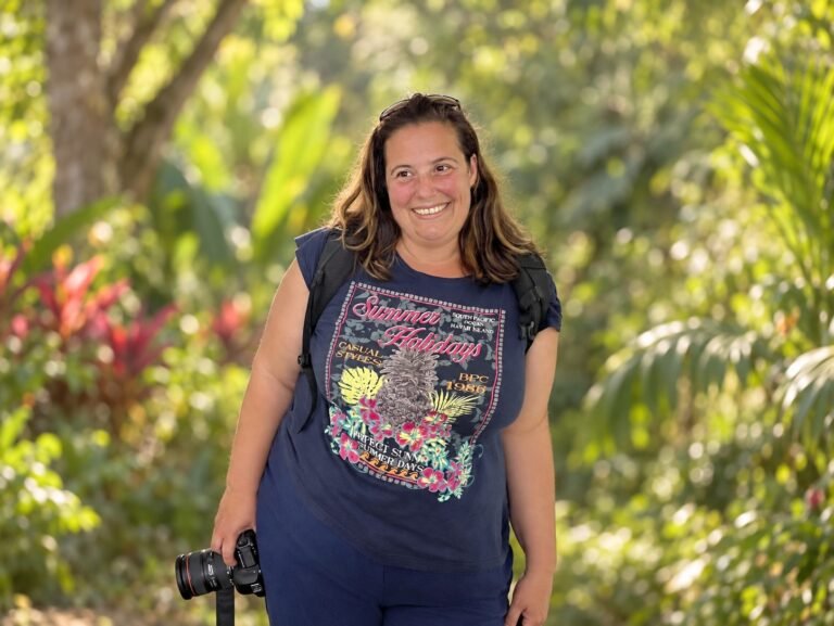 Fotografin Isabela Pacini lächelt mit einer Kamera in der Hand vor einer üppig grünen tropischen Gartenkulisse.