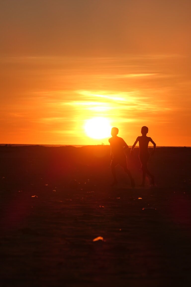 Zwei Kinder als dunkle Silhouetten vor der tief stehenden Sonne und einem leuchtend orangefarbenen Himmel.