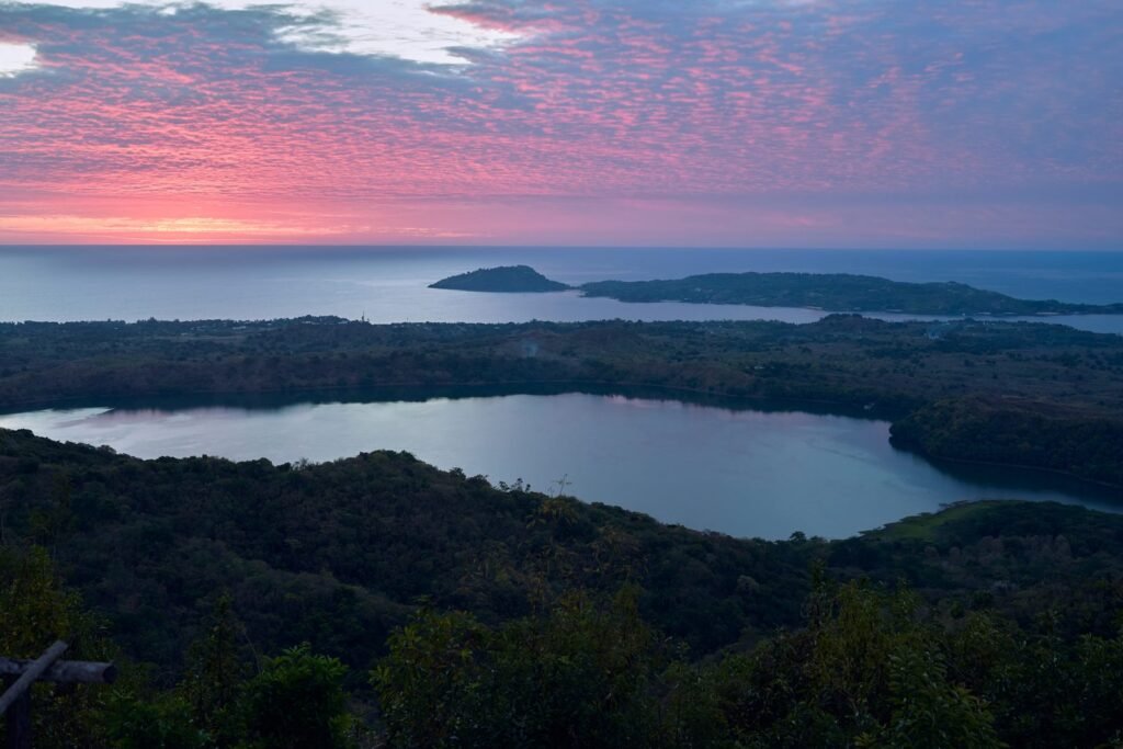 Blick vom Mont Passot auf Nosy Be über einen Kratersee und bewaldete Hügel zum Meer unter einem rosa leuchtenden Abendhimmel.